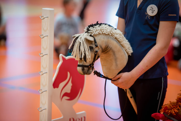 Close-up at the LarDen and CHHA indoor competition, with participants in navy sports shirts holding hobby horses beside a white show-jumping standard and red-and-white jump wing; a tan hobby horse with a cream-and-black yarn mane and bridle fills the
