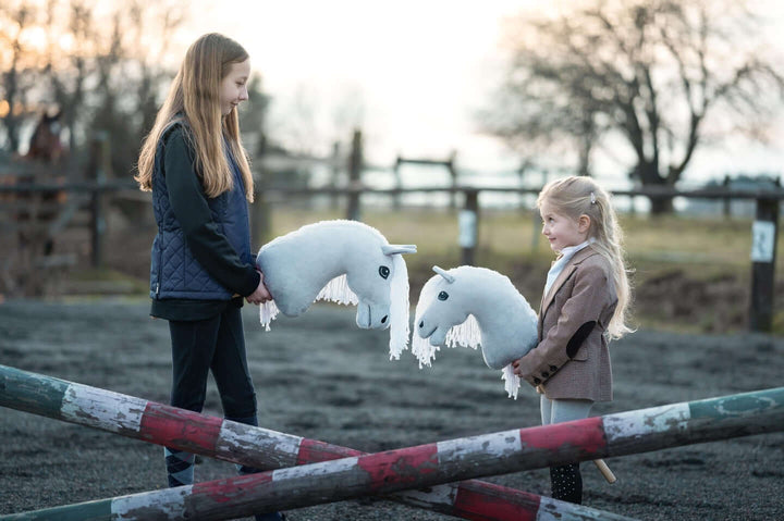 Dwoje dzieci trzymających białe Hobby Horses obok malowanych przeszkód na zewnętrznej arenie