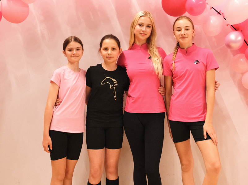 Four young riders stand arm in arm in front of a soft pink backdrop decorated with pink balloons, wearing sporty pink and black tops with black shorts or leggings, including hobby horse logo details. The bright, cheerful setup creates an inviting LarDen