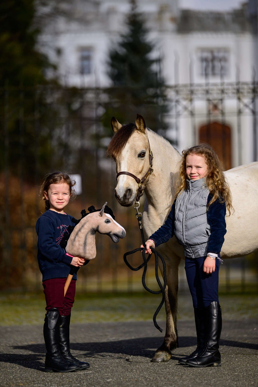 Dwie młode dziewczynki stoją na zewnątrz obok białego konia; jedna trzyma zabawkowego hobby horse, a druga wodze prawdziwego konia. W tle widać rozmyty budynek i drzewa.