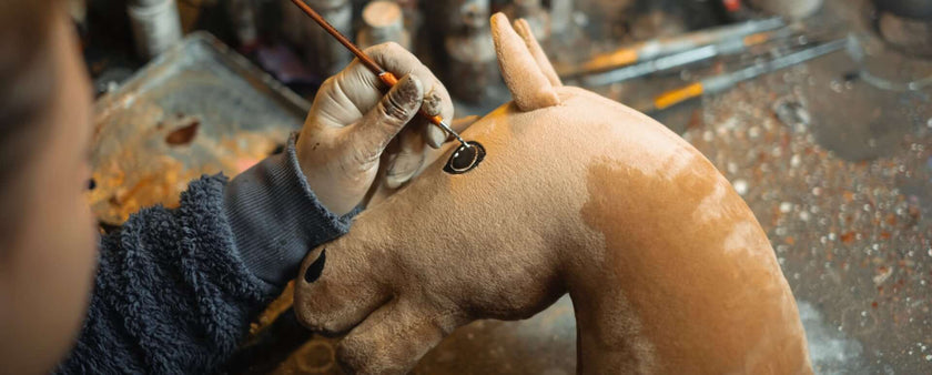 A person paints a black eye onto a brown Hobby Horse plush head, using a thin brush. The toy rests on a workbench scattered with art supplies and tools, capturing the creativity of hobby horsing enthusiasts.