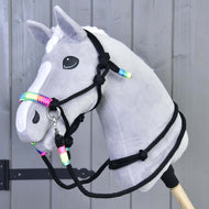 Szary pluszowy Hobby Horse od Hobby Horse LarDen z białą strzałką i grzywą, noszący kantar z zestawu Rope set Rainbow, osadzony na drewnianym kiju na tle szarych drewnianych paneli.