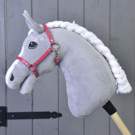 Szary pluszowy Hobby Horse LarDen z białą plecioną grzywą, czarnym okiem i Kantarem malinowym (rozmiar M), przymocowany do drewnianego kija, stoi przed szarą drewnianą ścianą z paneli.