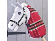Szara zabawka Hobby Horse od Hobby Horse LarDen, ubrana w Derkę stajenną Christmas w czerwoną kratę i czarną uzdę, jest osadzona na drewnianym kiju na tle jasnych drewnianych paneli.
