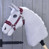 Kantar ze sztucznym futrem - skóra ekologiczna Burgundy od Hobby Horse LarDen, pokazany na szarym pluszowym Hobby Horse z białą grzywą i bordową uzdą, dodaje stylowego charakteru na tle szarej drewnianej ściany, czyniąc go idealnym dodatkiem.