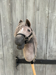 The Hobby Horse LarDen Hobby Horse Swing with Black Halter (size S) features a plush horse head on a wooden stick, displayed before a rustic gray fence.