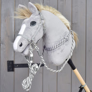 Zestaw Start Srebrny (Kantar + Cordeo + Uwiąz + wodze) od Hobby Horse LarDen na szarym pluszowym Hobby Horse z białą strzałką na pysku, osadzonym na drewnianym kiju i opartym o szarą drewnianą ścianę.