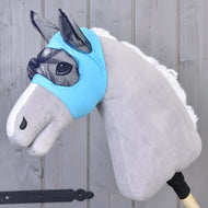A plush hobby horse with a light gray body, white mane, and a blue washable Hobby Horse LarDen Fly Mask, mounted on a wooden stick against a gray wooden background.
