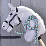 Hobby Horse - Pleciona lonża turkusowa od Hobby Horse LarDen przedstawia szarą pluszową głowę na drewnianym kiju, czarne ogłowie oraz plecioną linę w kolorach turkusowym, czerwonym i beżowym przewieszoną wokół szyi, na tle drewnianych paneli.