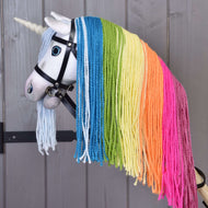 Hobby Horse Jednorożec Rainbow z Ogłowiem i Kantarem od Hobby Horse LarDen z białym rogiem, niebieskimi oczami, ogłowiem i wyrazistą tęczową grzywą z włóczki w kolorach niebieskim, zielonym, żółtym, pomarańczowym i różowym, na drewnianym tle.