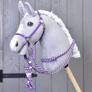 Pluszowy hobby horse z szarą głową, białą grzywą i białą strzałką ma założony Zestaw Start Fioletowy (kantar, cordeo, uwiąz, wodze) od Hobby Horse LarDen, osadzony na drewnianym kiju na tle szarej drewnianej ściany.