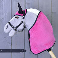 A gray stick horse toy by Hobby Horse LarDen, featuring a Set Hot Pink ear bonnet and blanket, with a black bridle, is positioned in front of a wooden fence.
