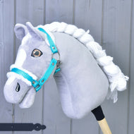 Szaro-biały Hobby Horse z plecioną białą grzywą nosi Kantar turkusowy z futerkiem Hobby Horse LarDen, idealne akcesorium, na tle jasnoszarego drewna.