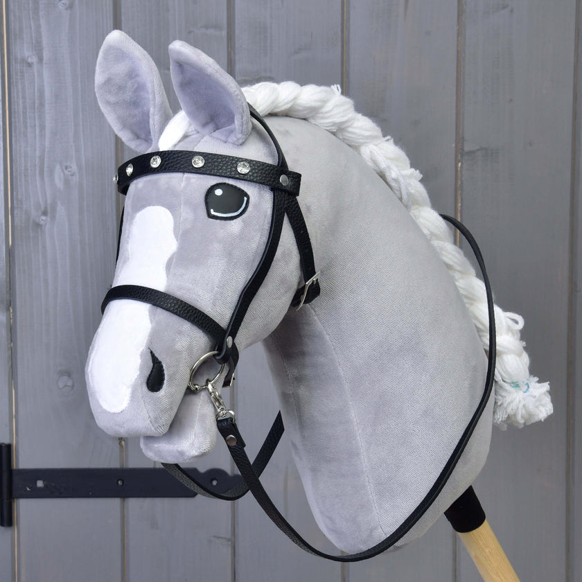 Szary pluszowy Hobby Horse od Hobby Horse LarDen ma kompletne czarne ogłowie z diamentami, białą grzywę, odpinane wodze i drewniany kij, ustawiony przed szarą drewnianą ścianą.