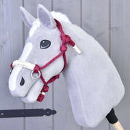 Szary pluszowy Hobby Horse od Hobby Horse LarDen z białą strzałką, czarnymi guzikowymi oczami i dodatkiem Sznurkowy Kantar Czi-Czi, osadzony na drewnianym kiju na tle szarej drewnianej ściany.