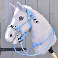 Pluszowy Hobby Horse z szarym futerkiem, białą grzywą i strzałką ma zestaw sznurkowy Malibu — niebieski kantar sznurkowy z czarno-białymi akcentami oraz pasujące wodze — osadzony na drewnianym kiju na tle szarego drewna.