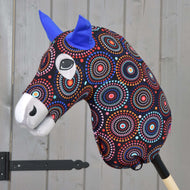 Hobby Horse LarDen Protective Suit Alice (Lycra) ma kolorową wzorzystą głowę, niebieskie filcowe uszy i wyraziste oczy na drewnianym kiju, pokazany na tle jasnoszarej drewnianej ściany.