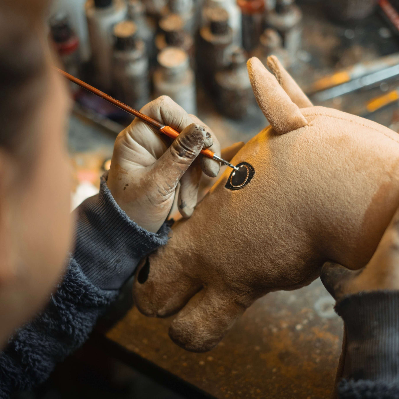 Osoba w rękawiczkach używa pędzelka, aby starannie malować oko brązowej pluszowej zabawki hobby horse, a w rozmytym tle widać słoiczki z farbą.