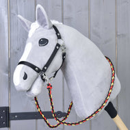 The Hobby Horse LarDen Reins Flag is a gray plush hobby horse on a wooden stick, with a black harness and handmade reins made of braided red, yellow, and black polypropylene rope, shown against a wooden wall.
