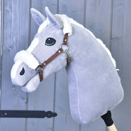 Pluszowy szary Hobby Horse Hobby Horse LarDen z białą grzywą i brązowym kantarem ze skóry ekologicznej z futrzanym wykończeniem, osadzony na drewnianym kiju na tle szarej drewnianej ściany.