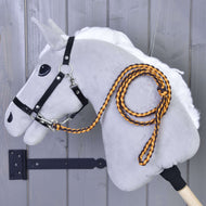 Hobby Horse - Dziana lonża Pomello od Hobby Horse LarDen przedstawia szarego pluszowego Hobby Horse z białą grzywą, czarną uzdą i pomarańczowo-czarną dzianą lonżą z karabińczykiem, zamocowanego na drewnianej ścianie.