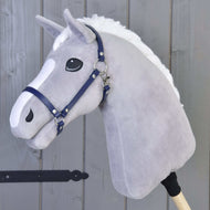 Szary pluszowy Hobby Horse z białą grzywą i kantarem Kantar - ciemnoniebieska skóra ekologiczna od Hobby Horse LarDen stoi na drewnianym kiju przed szarą drewnianą ścianą.