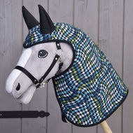 Pluszowy Hobby Horse LarDen w czarnym kantarze nosi płaszcz przeciwdeszczowy Lara z kapturem i uszami w niebiesko-zielono-biało-czarny wzór pepitki, pozując na tle drewnianego płotu.