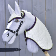 Zestaw Leonardo od Hobby Horse LarDen przedstawia szarego pluszowego Hobby Horse z białą pikowaną derką, pasującymi nausznikami, czarną uzdą i wodzami na drewnianym kiju, pokazany na tle szarej drewnianej ściany.