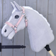 Hobby Horse LarDen Kantar ze sztucznym futrem - skóra ekologiczna Mullbery to jasnoszary pluszowy Hobby Horse z białą grzywą, różową uzdą ze skóry ekologicznej i haftowanymi detalami, osadzony na drewnianym kiju przed szarą drewnianą ścianą.