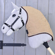 A close-up of the Hobby Horse LarDen Neck cover Bonanza—a gray hobby horse with black bridle and tan quilted blanket with secure fasteners—mounted on a wooden stick in front of a gray wooden wall.