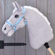 Szara pluszowa zabawka konia na kiju z białą grzywą ma założony Hobby Horse LarDen Kantar ze sztucznym futrem - skóra ekologiczna Pastell Blue i jest oparta o drewnianą panelową ścianę z czarnymi metalowymi zawiasami.