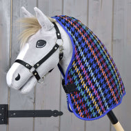 Biały zabawkowy Hobby Horse z czarnym kantarem ma na sobie kolorową derkę Derka stajenna Peru od Hobby Horse LarDen z wzorem pepitki, na tle jasnego drewna.
