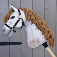 Zbliżenie na Hobby Horse Rufus od Hobby Horse LarDen z białym pyskiem, brązową grzywą z włóczki, czarnym kantarem i brązowymi cętkami na tle drewnianych paneli.