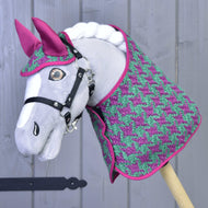 Szary Hobby Horse LarDen stoi przed jasnoszarą drewnianą ścianą, nosząc Zestaw Coco: różowo-zielony zestaw derek w pepitkę z dopasowanym różowym nausznikiem i czarną uzdą.