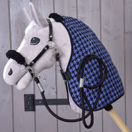 Pluszowy Hobby Horse Hobby Horse LarDen Set Harmony Blue z czarnym kantarem, wodzami i niebiesko-czarną derką stajenną w pepitkę jest zamocowany na ścianie na tle jasnych drewnianych desek.