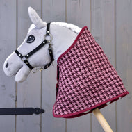 Hobby Horse LarDen „Zestaw świąteczny Bordeaux” przedstawia szarego pluszowego Hobby Horse z czarnym ogłowiem i bordowo-różową derką w pepitkę, osadzonego na drewnianym kiju jako urocza dekoracja na tle szarego drewna.