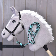 The Hobby Horse LarDen "Hobby Horse - Lead rope stripe green" features a plush gray horse with a white mane, black bridle, striped green lead rope, and is mounted on a wooden stick against a wooden wall.