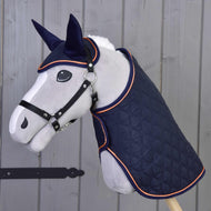 Zestaw Winner od Hobby Horse LarDen przedstawia pluszową głowę konia na patyku, ubraną w pikowaną granatową derkę i pasujący nausznik z pomarańczowym wykończeniem, na tle drewnianej ściany.