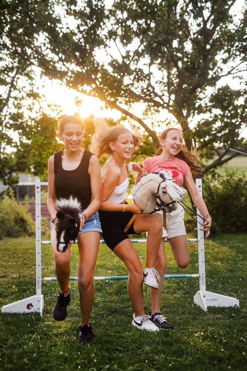 Trzy młode kobiety śmieją się, biegnąc i przeskakując przez przeszkodę na zewnątrz, każda trzymając Hobby Horse, a słońce prześwieca przez drzewa w tle, gdy cieszą się zabawą z Hobby Horsing.