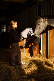 Ciemnowłosa dziewczynka w czarnej bluzie stoi w stajennym korytarzu, trzymając Hobby Horse Caesar z Black Halter od Hobby Horse LarDen, z szarą pluszową głową i czarno-białą włóczkową grzywą, gdy szary i gniady koń wyglądają z boksów, z ozdobnym ogłowiem bezwędzidłowym Complete