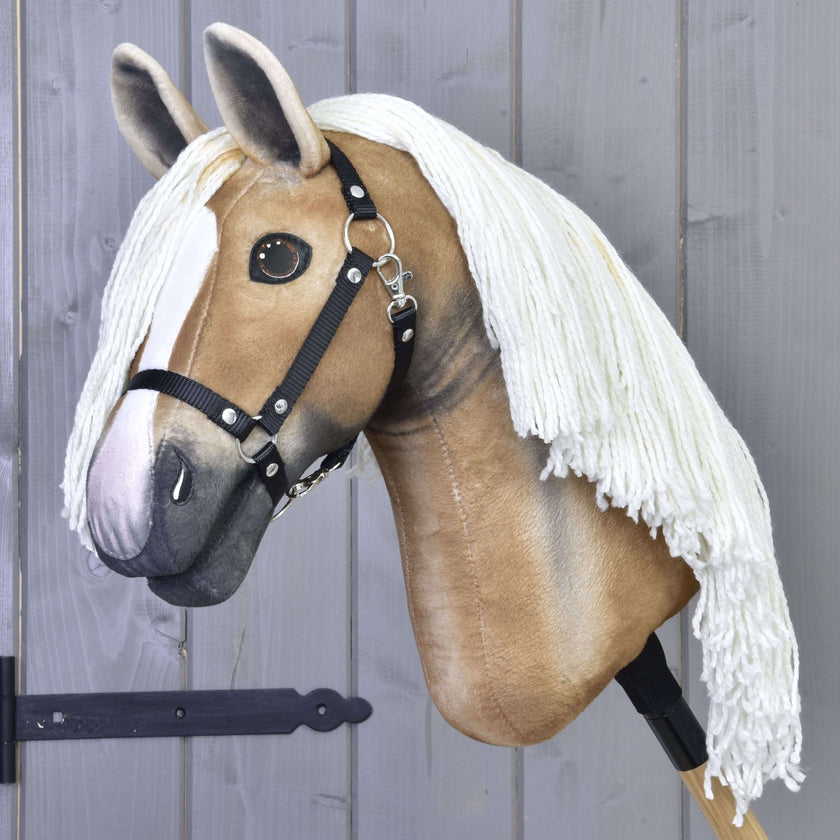 Hobby Horse Connie z Czarnym Kantarem od Hobby Horse LarDen ma realistyczny jasnobrązowo-biały wygląd, długą białą grzywę z włóczki i jest pokazany na kiju na tle szarej drewnianej ściany.