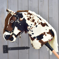 Hobby Horse Dakota z czarnym kantarem od Hobby Horse LarDen — pluszowy konik na kiju z brązowo-białymi cętkami, uzdą i grzywą z włóczki inspirowany dzikimi końmi rasy Mustang z Ameryki Północnej — pokazany na tle szarej drewnianej ściany.