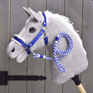 Zestaw Essential Niebieski. Hobby Horse LarDen Set Essential Blue przedstawia szarego pluszowego Hobby Horse z białą grzywą, niebieskim kantarem obszytym futerkiem i niebiesko-białą sznurkową wodzą na drewnianym kiju, pokazanym na tle szarego drewnianego płotu.