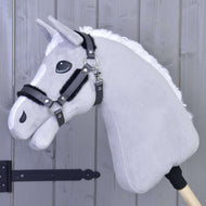 Kantar ze sztucznym futrem - skóra ekologiczna Graphite Full od Hobby Horse LarDen jest osadzony na drewnianym kiju i opiera się o jasnoszarą drewnianą ścianę; ma szarą pluszową głowę z białą grzywą, czarną uzdą i kantarem ze skóry ekologicznej.