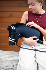 Dziewczyna w bordowej bluzce trzyma w ramionach Hobby Horse Mahal z kantarem Black Halter od Hobby Horse LarDen przed drewnianą ścianą – czarna pluszowa głowa, czarna grzywa z włóczki, biały futrzany Nachrapnik – pokazany z kompletnym Ogłowiem z futerkiem