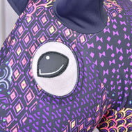 Close-up of the Hobby Horse LarDen Mandala protective suit (Lycra) showing its large embroidered eye and purple fabric with colorful geometric and diamond patterns; a dark ear appears at the top.