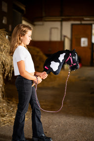Dziewczynka w białej bluzce prowadzi Hobby Horse Merlin z Czarnym Kantarem marki LarDen obok bel słomy w stajni – czarno-biała srokata pluszowa głowa, czarna grzywa z włóczki – pokazany z zestawem LarDen Exclusive Pink