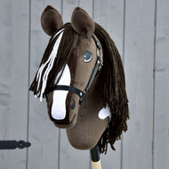 Hobby Horse LarDen's handmade Hobby Horse Monte features a brown and white coat inspired by the American Paint Horse, a black halter, long yarn mane, and is mounted on a wooden stick against a gray wood backdrop.