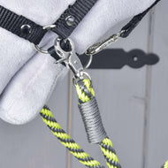 Close-up of a metal clasp attaching a black strap to the yellow, black, and gray "Hobby Horse - Reins lemon stripe" by Hobby Horse LarDen. The background is blurred gray wood with part of a soft white object visible.