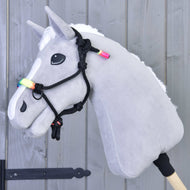Sznurkowy kantar Tęczowy Hobby Horse LarDen na szarym Hobby Horse z białą grzywą, tęczowymi koralikami i czarnym kantarem sznurkowym, osadzonym na drewnianym kiju, pokazanym na tle jasnoszarej drewnianej ściany.